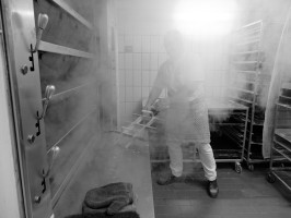 <span style='font-size:1.10em'>Ein Bäcker steht im Dampf einer gerade geschlossenen Ofenklappe.</span>