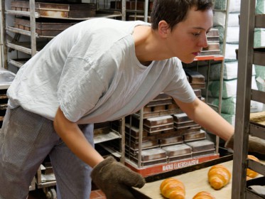 <span style='font-size:0.75em'><strong>Arbeiten am Ofen</strong> Die Bäckerin schiebt das Bleck mit den noch heißen Coissants zum Abkühlen in den Bäckereiwagen. ©Ernst Wilhelm Grüter</span>