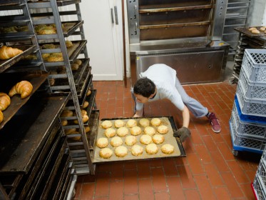 <span style='font-size:0.75em'><strong>Arbeiten am Ofen</strong> Die Bäckerin schiebt ein heißes Blech mit frisch gebackenen Käsebrötchen in den Bäckereiwagen. Sie unternimmt fast gymnastische Übungen, um die tiefste Ebene des Bäckereiwagens zu erreichen. ©Ernst Wilhelm Grüter</span>