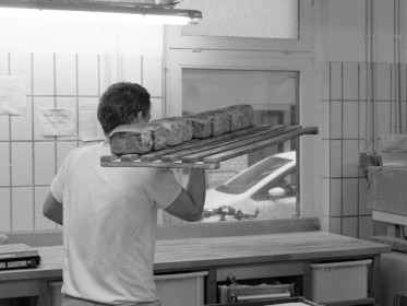 <span style='font-size:0.75em'><strong>Fertige Backwaren</strong> Ein Regalbrett mit Brotlaiben wird in das Ladengeschäft gebracht. Es liegt überm Hof. ©Ernst Wilhelm Grüter</span>