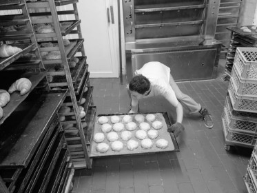 <span style='font-size:0.75em'><strong>Arbeiten am Ofen</strong> Die Bäckerin schiebt ein heißes Blech mit frisch gebackenen Käsebrötchen in den Bäckereiwagen. Sie unternimmt fast gymnastische Übungen, um die tiefste Ebene des Bäckereiwagens zu erreichen. ©Ernst Wilhelm Grüter</span>